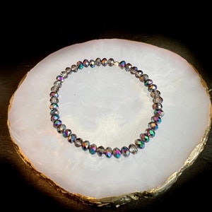 May include: A beaded bracelet with iridescent, faceted beads. The beads are primarily gray with flashes of green, blue, and purple. The bracelet is displayed on a white, oval-shaped surface with a gold-colored edge.