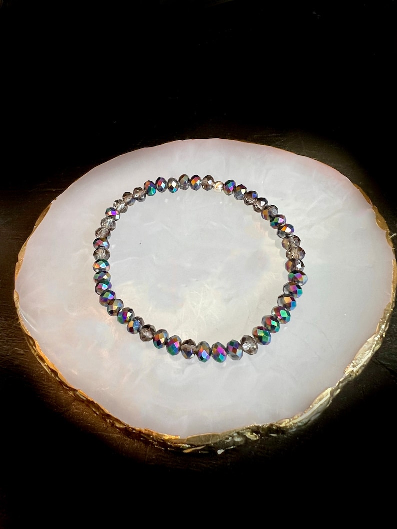 May include: A beaded bracelet with iridescent, faceted beads. The beads are primarily gray with flashes of green, blue, and purple. The bracelet is displayed on a white, oval-shaped surface with a gold-colored edge.