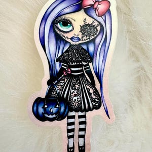 May include: A sticker featuring a gothic doll with blue hair and a pink bow. The doll wears a black and white striped dress and holds a blue jack-o'-lantern. The doll's face has stitched details and a decorative eye patch.
