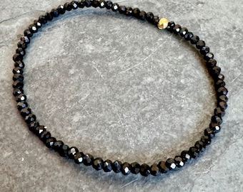 May include: A delicate black beaded bracelet with a small gold accent bead. The bracelet is made of small, faceted black beads strung together, creating a subtle sparkle. The gold bead adds a touch of contrast and elegance to the design.