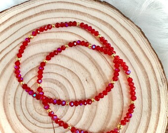 May include: Two red beaded bracelets with gold-colored accents are displayed on a wooden surface. The beads have a faceted, crystal-like appearance, reflecting light. The bracelets are arranged in a figure-eight pattern.
