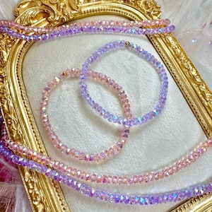May include: Two beaded bracelets and two strands of beads in shades of pink and purple. The bracelets are circular, with small, faceted beads. The strands of beads are laid out in a decorative pattern. The background is a gold-colored frame.