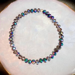 May include: A beaded bracelet with iridescent, faceted beads in shades of gray, purple, and green. The bracelet is circular and sits on a white, oval-shaped surface.