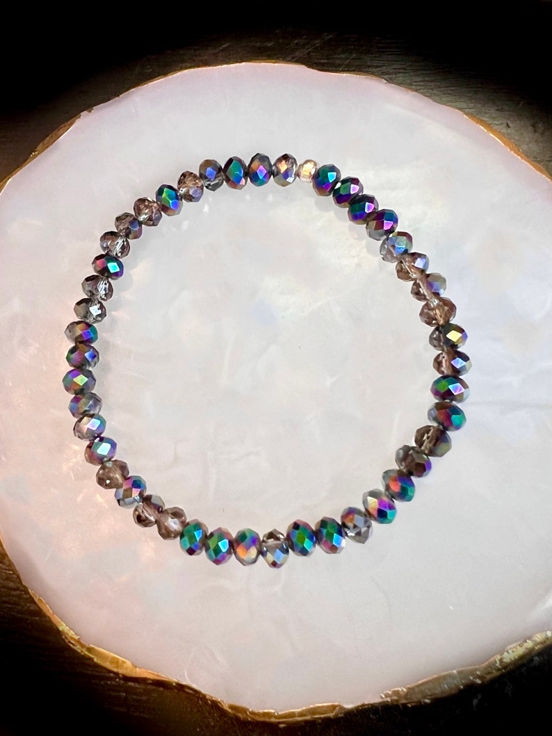May include: A beaded bracelet with iridescent, faceted beads in shades of gray, purple, and green. The bracelet is circular and sits on a white, oval-shaped surface.