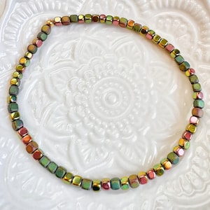 May include: A beaded bracelet featuring small, square beads in shades of green, gold, and pink. The bracelet is strung together with a gold-colored clasp. The beads have a metallic sheen, giving the bracelet a unique look.