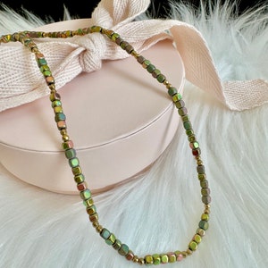 May include: A delicate necklace featuring small, square beads in shades of green, gold, and brown. The beads are strung together with gold-colored spacers. The necklace is displayed on a pink box with a cream-colored ribbon.