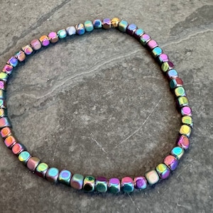 May include: A beaded bracelet with iridescent, cube-shaped beads. The beads display a spectrum of colors, including purple, green, blue, and gold. The bracelet is strung on a flexible cord, creating a delicate and eye-catching accessory.