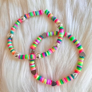 May include: Two colorful beaded bracelets with a rainbow of colors, including pink, green, yellow, and purple. The bracelets feature silver beads and heart-shaped charms. The bracelets are displayed on a white, fluffy surface.