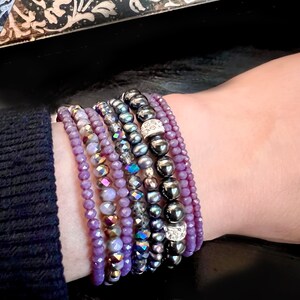 May include: A collection of beaded bracelets in shades of purple, gray, and black. The bracelets feature various bead sizes and textures, including faceted beads, smooth spheres, and beads with iridescent and rhinestone accents. The bracelets are stacked on a wrist.