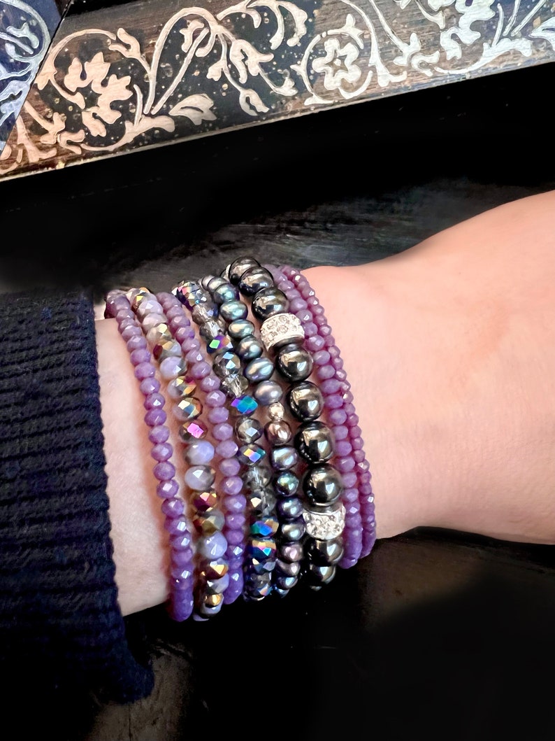 May include: A collection of beaded bracelets in shades of purple, gray, and black. The bracelets feature various bead sizes and textures, including faceted beads, smooth spheres, and beads with iridescent and rhinestone accents. The bracelets are stacked on a wrist.