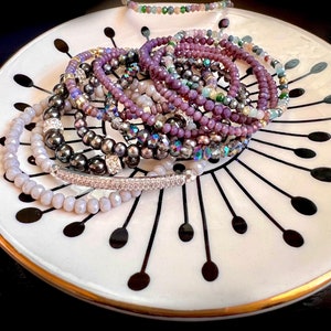 May include: A collection of beaded bracelets in various shades of purple, gray, and iridescent colors. The bracelets are displayed on a white plate with a black and gold design. The bracelets are made of different materials, including faceted beads and pearls.