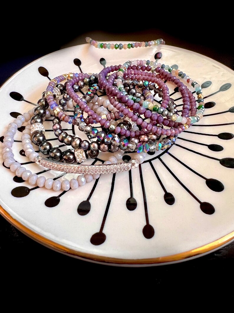 May include: A collection of beaded bracelets in various shades of purple, gray, and iridescent colors. The bracelets are displayed on a white plate with a black and gold design. The bracelets are made of different materials, including faceted beads and pearls.