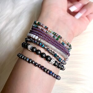 May include: A collection of beaded bracelets in various colors and styles. The bracelets include dark gray, purple, and iridescent beads, with some featuring silver accents. The bracelets are stacked on a wrist against a white, fluffy background.