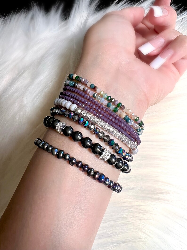 May include: A collection of beaded bracelets in various colors and styles. The bracelets include dark gray, purple, and iridescent beads, with some featuring silver accents. The bracelets are stacked on a wrist against a white, fluffy background.