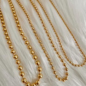 May include: Four gold-colored ball chain necklaces of varying lengths are displayed against a white, fluffy background. The necklaces are made of small, round beads strung together, creating a delicate and elegant look. The chains are arranged to showcase their different sizes.