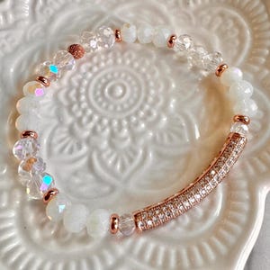 May include: A white beaded bracelet with clear and iridescent beads, accented with rose gold-colored metal spacers and a curved bar encrusted with small, clear crystals. The bracelet is displayed on a white, patterned surface.