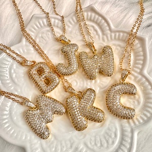 May include: Gold-toned necklaces with initial pendants. Each pendant is a letter of the alphabet, encrusted with small, sparkling crystals. The necklaces are displayed on a white, decorative plate.