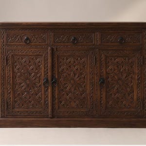 May include: A dark brown wooden sideboard featuring three top drawers and three lower cabinet doors. The front panels are decorated with intricate floral carvings and geometric borders, complemented by circular metal ring pulls on each drawer and door.