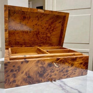 Peut inclure: Coffret en bois avec couvercle à charnière, ouvert pour révéler un intérieur avec compartiments. Le coffret est en bois aux tons chauds avec un motif de grain distinctif. Un petit trou de serrure est visible à l'avant.
