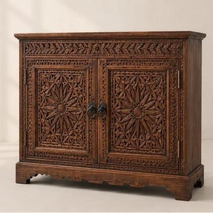 Peut inclure: Un meuble rectangulaire en bois brun foncé doté de deux portes ornées de motifs sculptés à la main en forme d'étoiles et de feuilles. Le meuble comprend deux poignées en anneau en métal noir au centre des portes et repose sur quatre pieds sculptés.