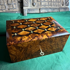 Peut inclure: Une boîte à bijoux rectangulaire en bois avec un dessus à motifs. La boîte est faite de bois sombre et veiné avec une finition brillante. Le dessus présente un motif en losanges avec des incrustations. Une petite clé est dans la serrure.