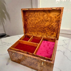 May include: An open wooden jewelry box with a burl wood exterior and a red velvet interior. The box is divided into compartments, with a pink velvet lining in one section. The box is sitting on a white marble table.