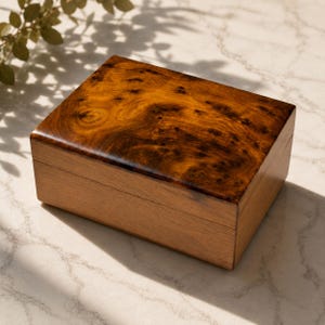 Peut inclure: Une boîte rectangulaire en bois avec un couvercle à charnière présentant un motif de grain de bois de ronce brun foncé poli sur le dessus. Les côtés de la boîte sont faits d'un bois brun clair uni avec une texture droite et une finition lisse.