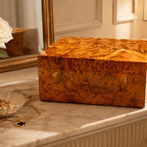 May include: A rectangular wooden box with a polished, high-gloss finish featuring a distinct, swirling burl wood grain pattern in shades of amber and brown. The box has a hinged lid and two small, metallic gold-toned latches on the front panel.
