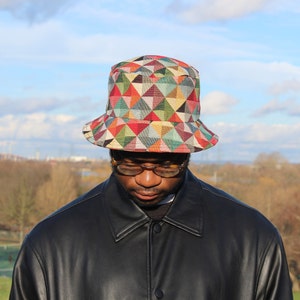 May include: A colorful geometric patterned bucket hat with red, orange, green, and blue triangles. The hat is worn by a person wearing glasses and a black leather jacket. The background features a park and a cloudy sky.