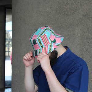 May include: A colorful bucket hat with a geometric pattern in pink, teal, green, and red. The hat is worn with a blue shirt. The hat's design features a patchwork of shapes and lines, creating a vibrant accessory.