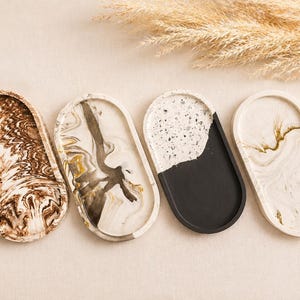 May include: Four oval decorative trays with unique patterns. The trays feature marbled effects in brown, black, and white, and a speckled black and white design. The trays are arranged on a neutral surface.