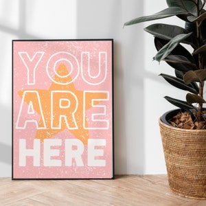 Puede incluir: Una impresión enmarcada rosa y blanca con el texto "YOU ARE HERE" en letras blancas y una gran estrella naranja en el centro.