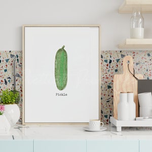 May include: A framed print of a green pickle with the word "Pickle" printed below it. The print is hanging on a wall in a kitchen.