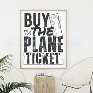 May include: A framed print with the words "BUY THE PLANE TICKET" in a bold, distressed black font. The design includes a paper airplane and a ticket illustration, all on a white background. The print is in a light wood frame.