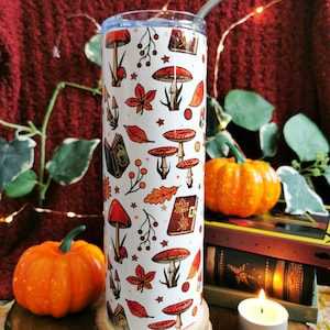 May include: A white stainless steel tumbler with a fall-themed design featuring red mushrooms, orange leaves, and brown books. The tumbler has a straw and is sitting on a wooden coaster.