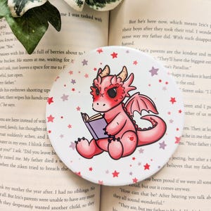 May include: A pink dragon with red eyes is sitting on a white background with pink and purple stars. The dragon is holding a purple book. The text "They'll have the biggest estate in Na's Paradise." is visible in the background.
