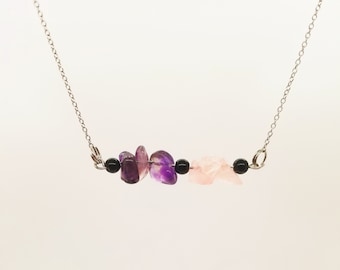 May include: A silver chain necklace featuring a horizontal bar with amethyst, rose quartz, and black bead accents. The gemstones are in shades of purple and pink, creating a delicate and elegant design. The necklace is displayed against a white background.