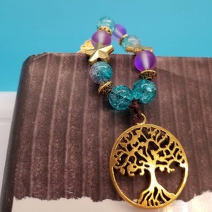 May include: A beaded charm with a gold-colored tree of life pendant. The charm features turquoise, purple, and clear beads, along with a gold star-shaped charm. The beads are strung on a brown cord.