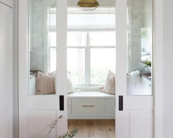 May include: Interior shot of a bright, airy space with white sliding doors and a view of a window seat. A woven basket with greenery sits on the wooden floor. A brass pendant light hangs above the window.