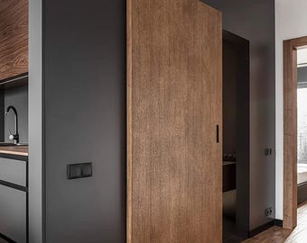 May include: A tall, rectangular sliding door made of medium-toned wood with a visible grain pattern, mounted against a matte charcoal gray wall. The door features a vertical black recessed handle and is positioned next to a dark kitchen cabinet.