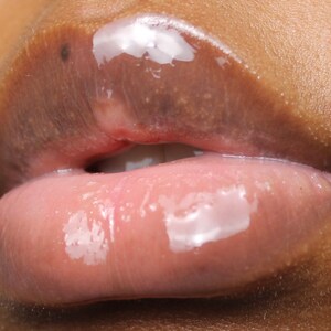 May include: Close-up of a person's lips wearing clear lip gloss with a pink tint. The gloss is shiny and has a wet look.