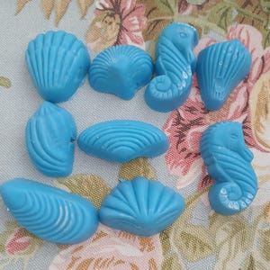 May include: A collection of eight bright blue wax melts shaped like various sea creatures and shells, including two seahorses, four fan-shaped shells, one elongated spiral shell, and one smooth, rounded shell, all arranged in a loose cluster.