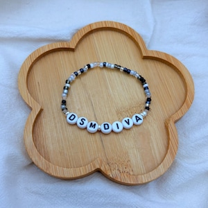 May include: A beaded bracelet with the word "DSMDIVA" spelled out in white letter beads. The bracelet features a mix of black, clear, and silver beads. It rests on a wooden, flower-shaped tray.