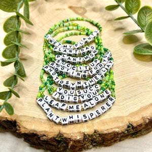 Könnte beinhalten: Eine Sammlung von Armbändern aus grünen und weißen Perlen. Jedes Armband hat weiße Buchstabenperlen, die pflanzliche Sätze wie "Plant Dad" und "Leaf Me Alone" buchstabieren. Die Armbänder liegen auf einer Holzoberfläche.