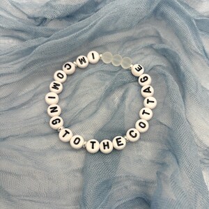 May include: A white beaded bracelet with black letter beads spelling "COMING TO THE COTTAGE." The bracelet also features clear, frosted beads. The bracelet is displayed on a light blue, textured fabric.
