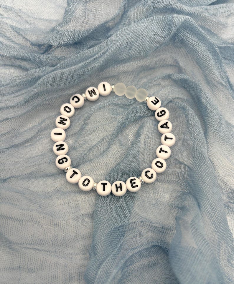 May include: A white beaded bracelet with black letter beads spelling "COMING TO THE COTTAGE." The bracelet also features clear, frosted beads. The bracelet is displayed on a light blue, textured fabric.