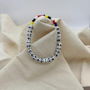 May include: A beaded bracelet with white letter beads spelling "CANADIAN STUPID BIRD WOLF WOMAN". The bracelet has colorful beads in red, blue, yellow, and black. The bracelet is on a beige fabric.