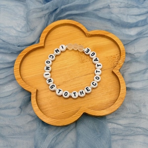 May include: A beaded bracelet with white lettered beads spelling "COMING TO THE COTTAGE" is displayed on a wooden, flower-shaped tray. The bracelet has clear beads and is set against a blue fabric backdrop.