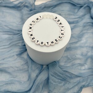 May include: A white beaded bracelet with black letter beads spelling "I'M COMING TO THE COTTAGE" is displayed on a white cylindrical stand. The bracelet has clear beads and is set against a blue fabric backdrop.