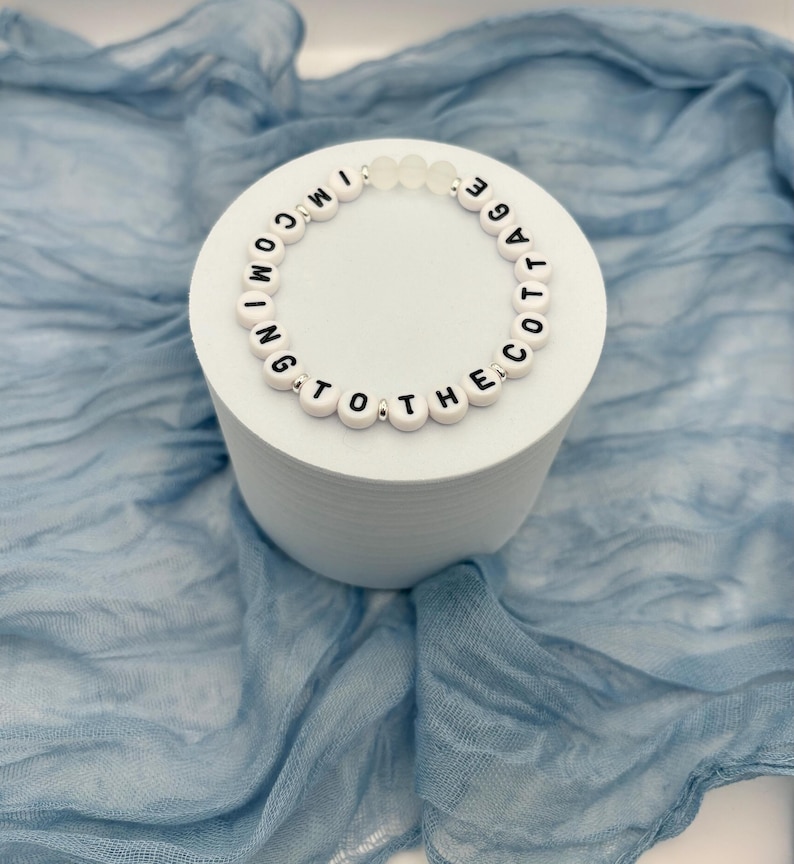 May include: A white beaded bracelet with black letter beads spelling "I'M COMING TO THE COTTAGE" is displayed on a white cylindrical stand. The bracelet has clear beads and is set against a blue fabric backdrop.